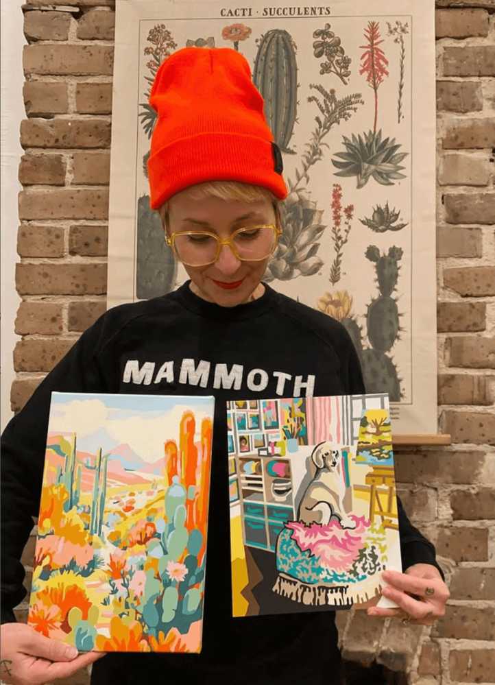 A lady showing off her 2 paint by numbers finished paintings.