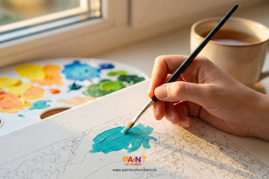 Close-up of a hand painting a teal section on a paint-by-numbers canvas with a palette and warm light coming through the window.