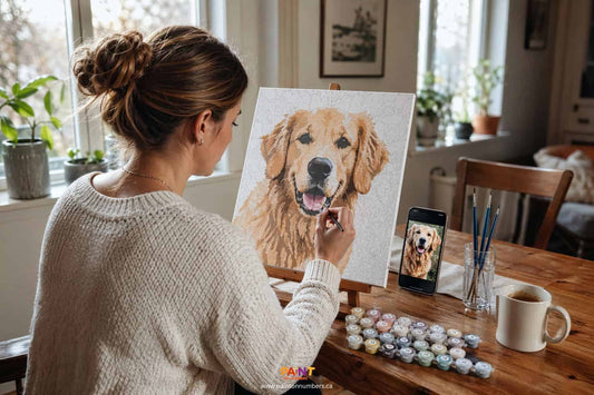 A Canadian woman sitting at a wooden table painting a custom paint by numbers portrait of her golden retriever, with the original photo visible on her phone beside the canvas