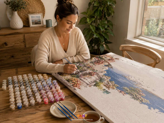 Woman working on a paint by numbers project with a scenic design, surrounded by paint pots, brushed and a cup of coffee.