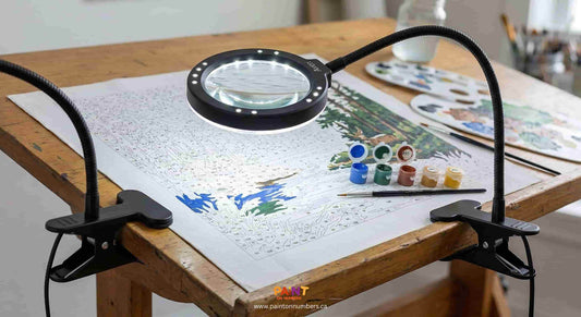 Adjustable clip on LED magnifying lamp illuminating a detailed paint by numbers canvas on a wooden drafting table