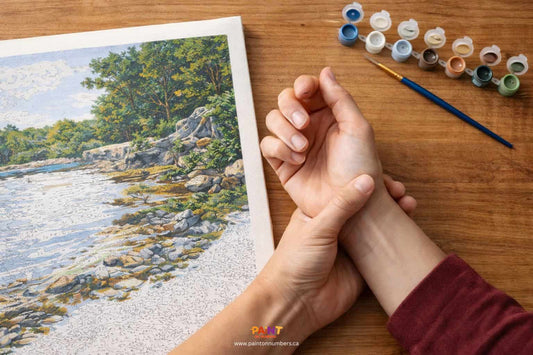 A person gently massaging their right wrist due to cramping next to a paint by numbers canvas and brushes on a wooden table.