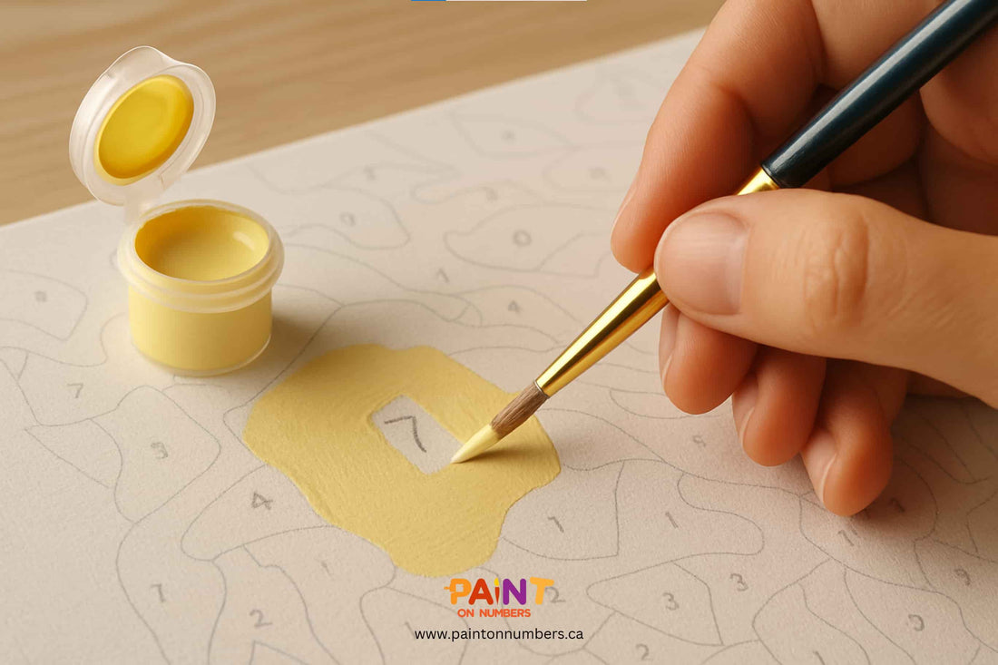 Close-up of a hand applying a second coat of yellow paint on a paint by numbers canvas to cover the printed number, with brush and paint pot in view.