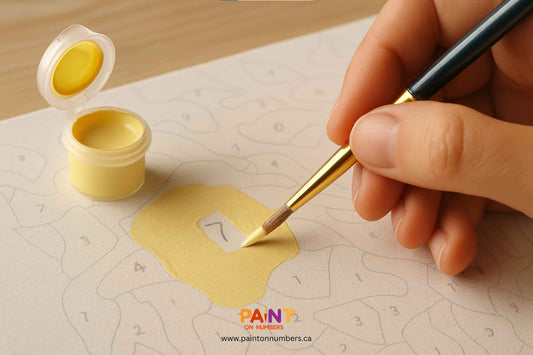 Close-up of a hand applying a second coat of yellow paint on a paint by numbers canvas to cover the printed number, with brush and paint pot in view.