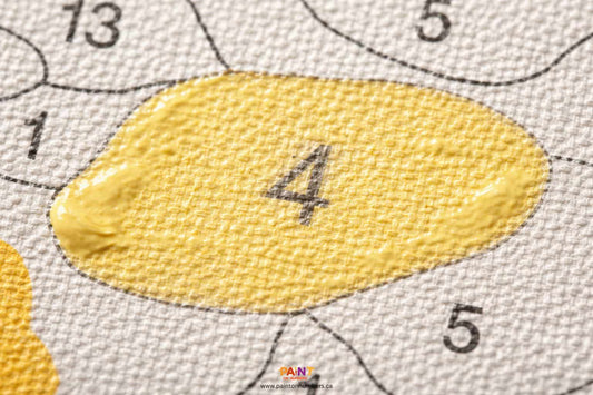 A close up of a paint by numbers canvas showing a light yellow painted cell where the dark grey number is still faintly visible underneath.