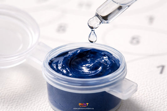 A top-down close up of a glass dropper adding a single drop of distilled water into a small pot of thick blue acrylic paint.