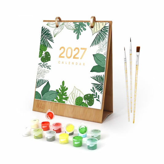 A 2027 desk calendar with a bamboo stand, decorated with green tropical leaves and gold text, shown with a set of small paint pots and three paintbrushes on a white background.
