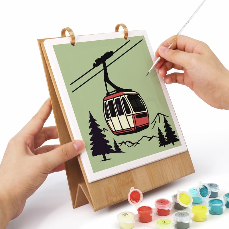 A diamond painting kit in progress, featuring a bamboo stand holding a canvas with a cable car illustration. Hands are shown painting over the illustration, with paint pots arranged on the white surface below.