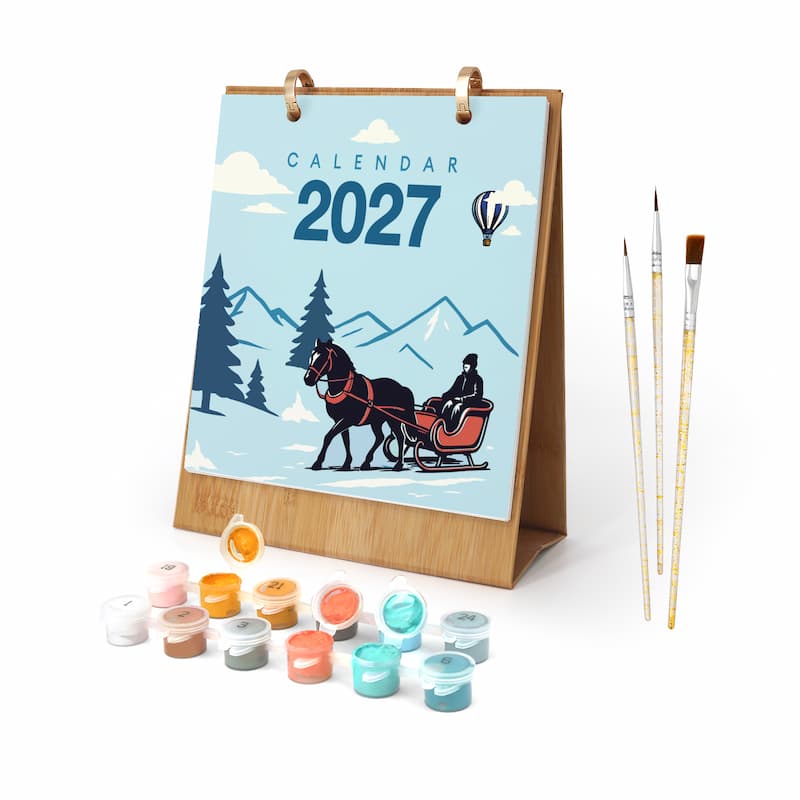 A 2027 desk calendar with a winter scene illustration, shown with paint pots and three paintbrushes on a white background.