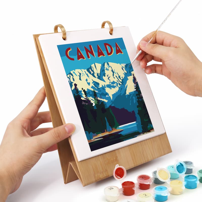 A person's hands are shown painting a travel poster of Canada on a bamboo A-frame easel. The poster features a colorful illustration of snow-capped mountains, pine trees, and a lake under a blue sky with 'CANADA' written in red letters.