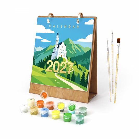A 2027 landscape calendar painting set on a wooden easel, featuring a colorful illustration of a white castle in a green mountain valley under a blue sky. The set includes three paintbrushes and multiple small pots of acrylic paint in various colors arranged in front of the easel.