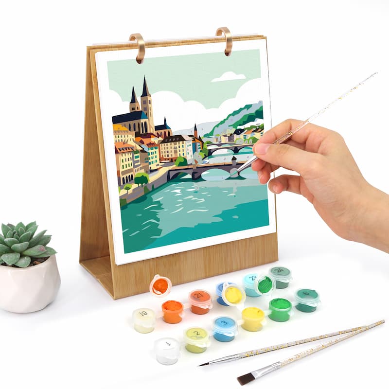 A hand holds a fine paintbrush next to a wooden stand holding a colorful cityscape painting kit. Small paint pots and two paintbrushes are arranged in front on a white surface, with a small potted succulent to the left.