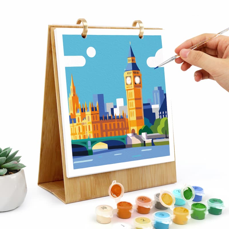 A wooden stand-mounted painting kit shows a colorful illustration of Big Ben and London landmarks being painted by a person's hand holding a fine brush. Several small paint pots are arranged in front on a white background.