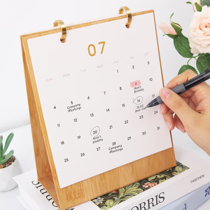 A bamboo desktop calendar with a white page showing the 7th of a month, held by a hand with a pen, styled on top of books with a rose in the background.