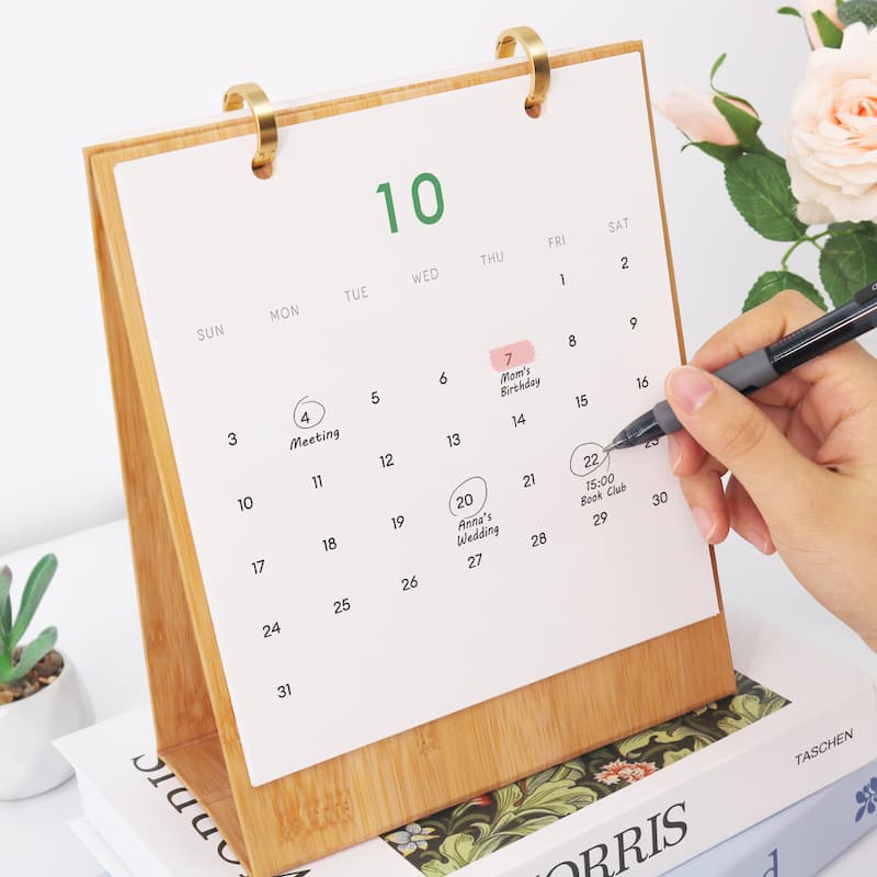 A bamboo desk calendar with a white page showing the 10th and a hand writing on it, styled on books with a rose in the background.