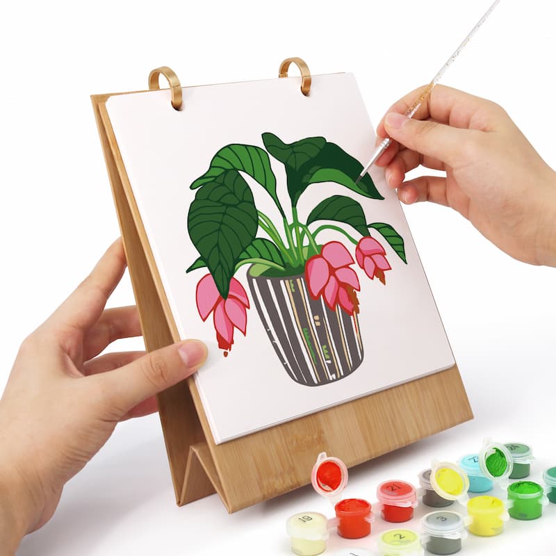 A person holds a wooden stand holding a canvas with a partially painted illustration of a potted plant with pink flowers. Small paint pots are arranged at the bottom right on a white background.