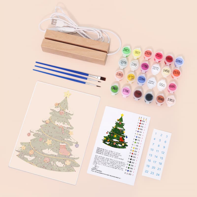 A Christmas tree painting kit laid out on a light beige background, including a wooden stand, USB charging cable, paintbrushes, multiple small paint pots, a printed Christmas tree design sheet, instruction card, and numbered sticker sheet.