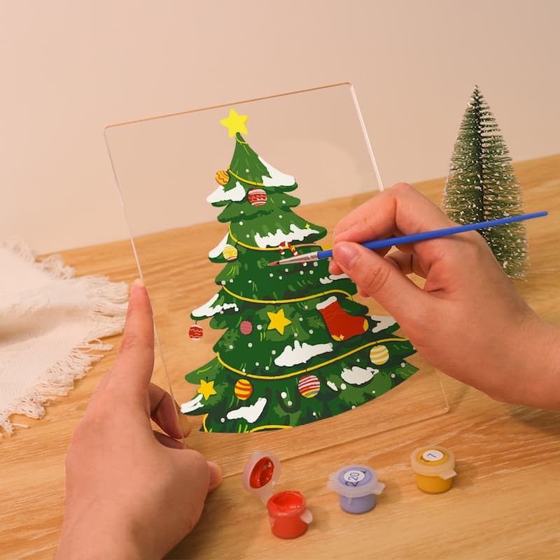 Hand painting a Christmas tree on a clear acrylic panel with small containers of paint on a wooden surface.