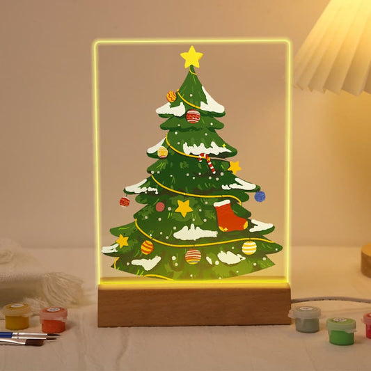 A glowing acrylic LED light box shaped like a Christmas tree, displayed on a wooden base on a tabletop. The tree illustration is decorated with snow, stars, ornaments, and a red stocking, surrounded by warm yellow light.