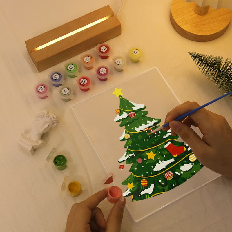 A person's hands are painting a Christmas tree with acrylic paints on a clear acrylic panel, surrounded by a wooden light box, paint pots, and a small decorative tree on a light wood surface.
