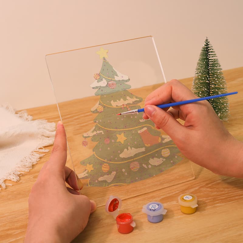 A person's hands are shown painting a clear acrylic panel with a Christmas tree design, using a fine paintbrush. Small paint pots are visible on a wooden table next to a miniature Christmas tree.