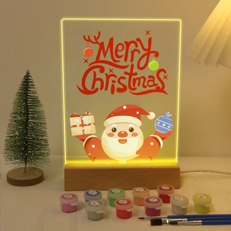 A glowing acrylic LED sign with a Christmas design, featuring the words 'Merry Christmas' in red script and a cartoon Santa holding gifts and an ornament. The sign is displayed on a wooden base on a tabletop, with a small Christmas tree, paint pots, and paintbrushes nearby.