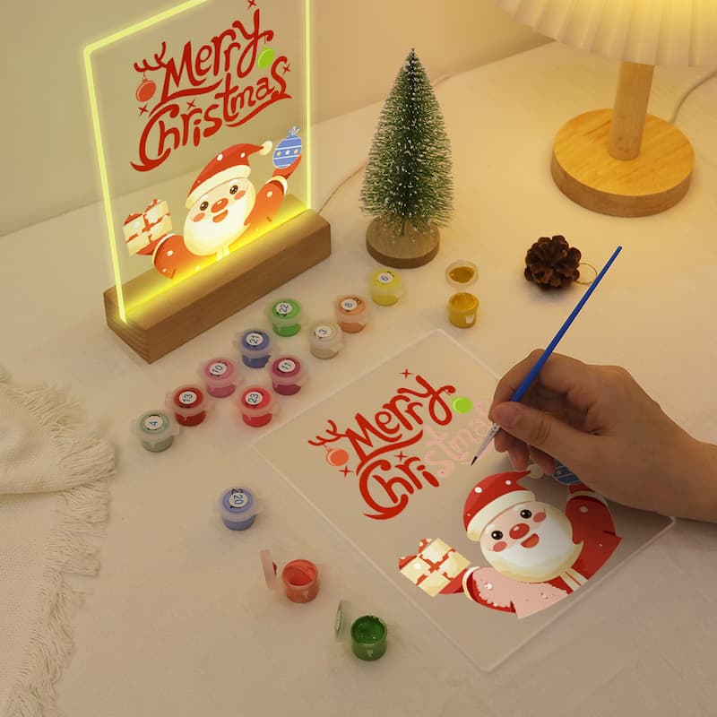 Decorative Christmas scene with a hand painting a card, a lit 'Merry Christmas' sign, and small Christmas tree.