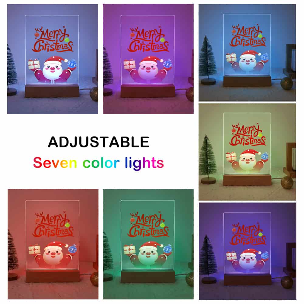 A collage showing a tabletop Christmas light in seven different color modes, with the text 'ADJUSTABLE Seven color lights' in the center. The light features a clear panel displaying a cartoon Santa and 'Merry Christmas' lettering, mounted on a wooden base and shown with small decorative trees.