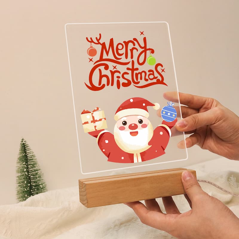A clear acrylic LED light box displaying a festive Christmas design with the words 'Merry Christmas' in red and a cartoon Santa holding gifts, resting on a wooden base and held by two hands.
