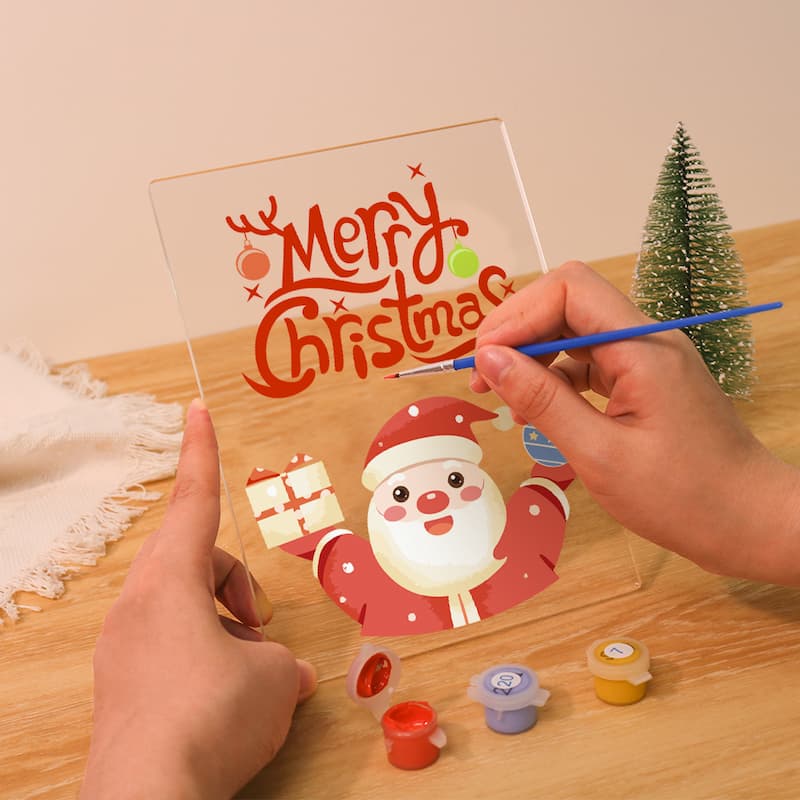Person painting a Christmas-themed acrylic block with a Santa Claus design on a wooden table.