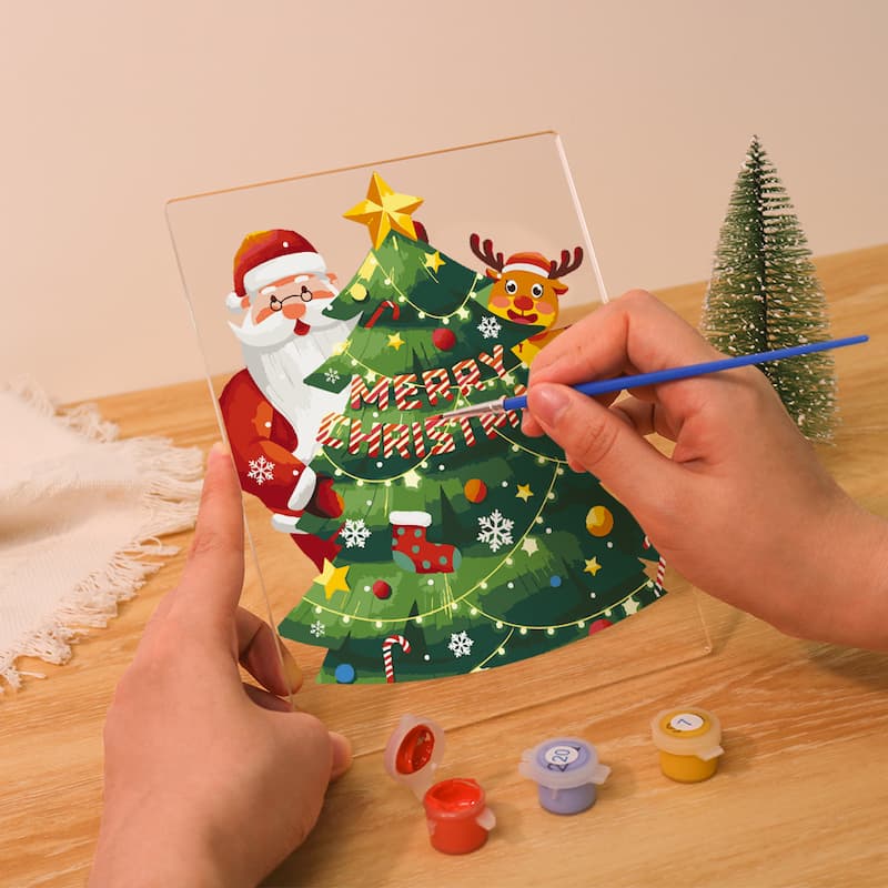 A person's hands are shown painting a clear acrylic panel with a Christmas tree design. The panel features Santa Claus, a reindeer, a green Christmas tree with colorful ornaments, and 'MERRY CHRISTMAS' text.