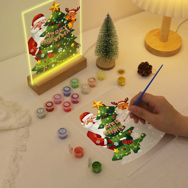 A hand is painting a Christmas tree and Santa design on a clear panel, with small paint pots arranged nearby on a tabletop. A glowing framed Christmas light and a small tree lamp are also visible in the background, creating a cozy holiday scene.