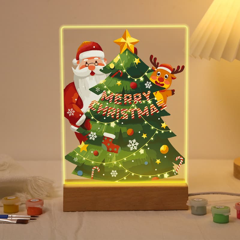 A rectangular LED light box on a wooden base featuring a festive Christmas illustration with Santa Claus, a reindeer, a decorated tree, and the words “Merry Christmas,” glowing with a warm yellow light in a cozy indoor setting.