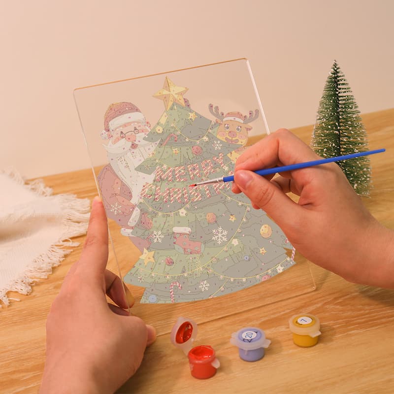 Person holding a decorative card with Santa Claus and a Christmas tree, using a paintbrush on a wooden surface.