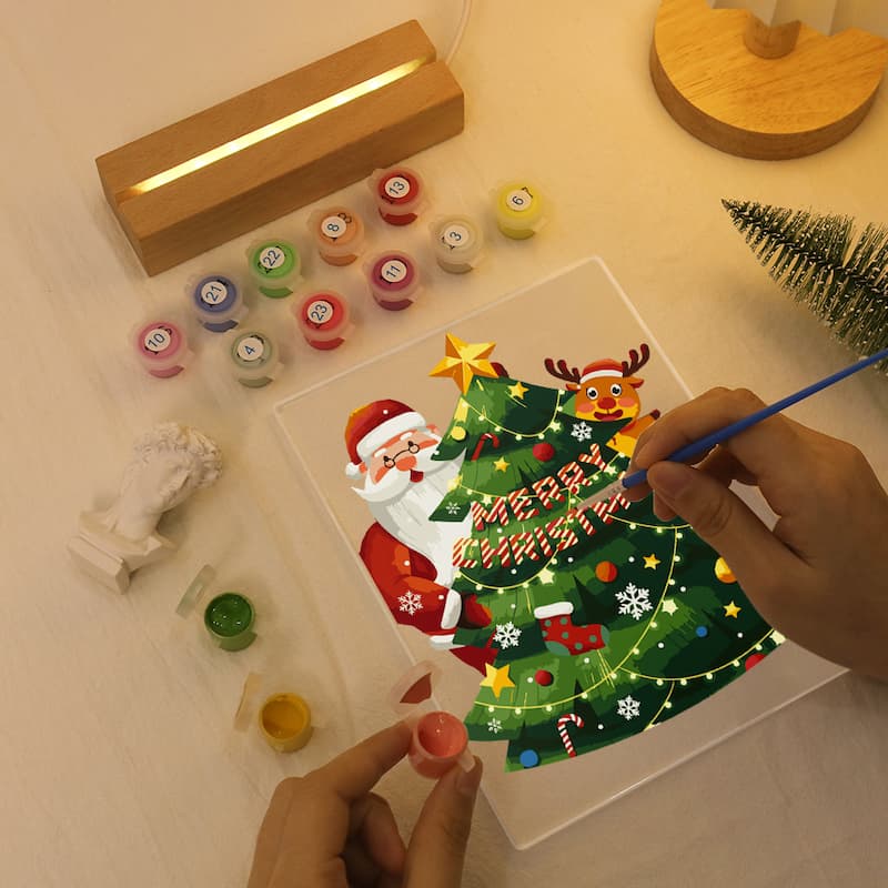 A Christmas-themed diamond painting kit in use, with hands painting a Santa Claus and tree design on a clear panel. The scene includes small paint pots, a wooden light box, a miniature tree, and other craft pieces on a tabletop.