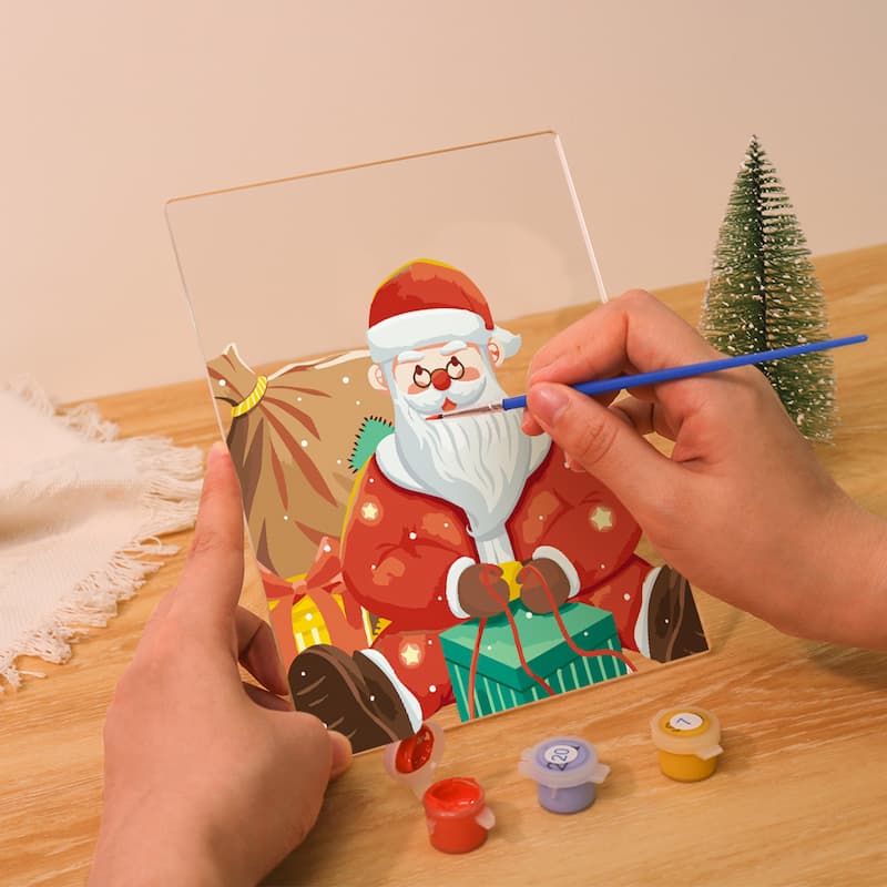 Hand painting a Christmas-themed picture of Santa Claus on a clear acrylic panel with small containers of paint.