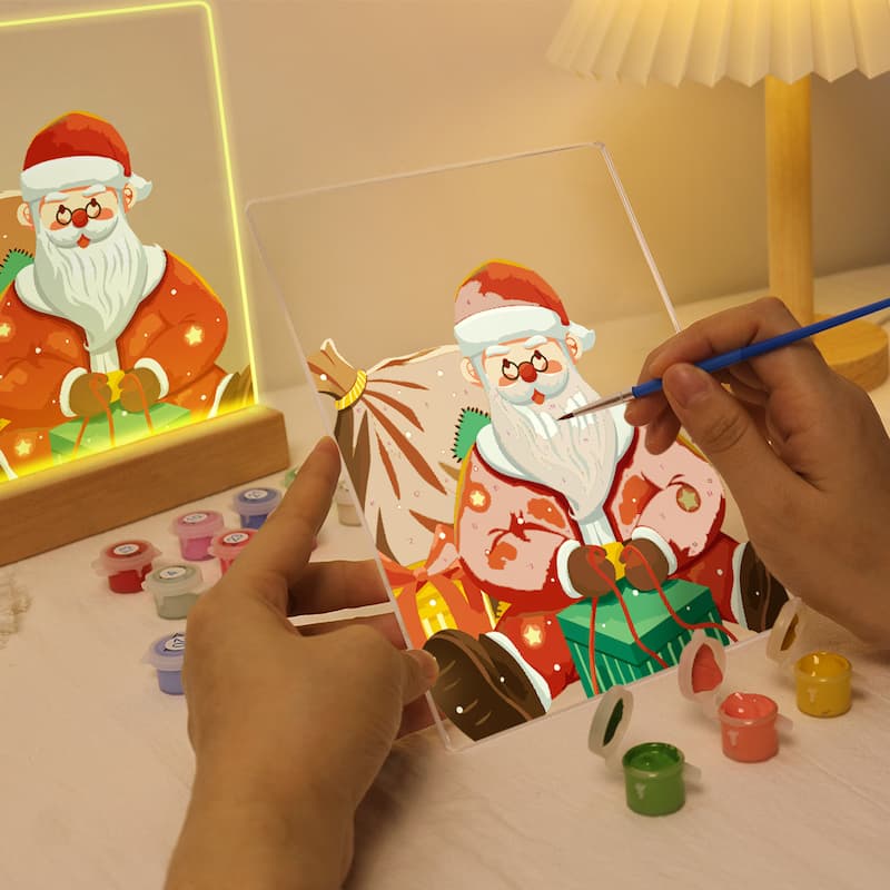 A person's hands are shown painting a Christmas-themed canvas with Santa Claus, while a lit LED light-up canvas of Santa Claus sits in the background on a wooden stand. Small paint pots are scattered on the table.