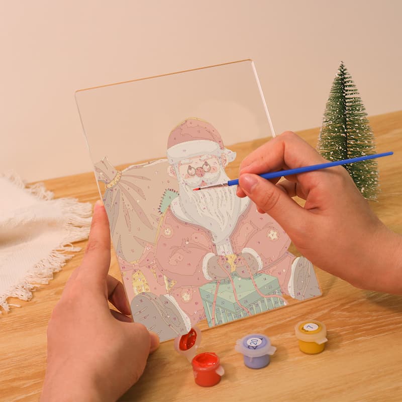 A person holds a clear acrylic panel with a colorful Santa Claus illustration while painting over it with a fine-tip brush. Small paint pots sit on a wooden table beside a miniature Christmas tree.