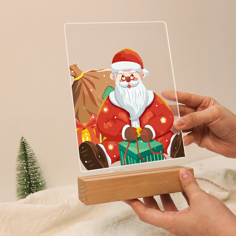 A hand holds a clear acrylic plaque featuring a colorful illustration of Santa Claus sitting with gifts and a sack, mounted on a light wood base. The other hand supports the base.