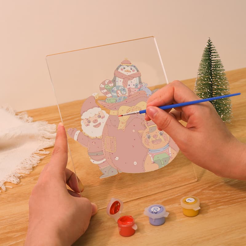 Person painting a transparent acrylic panel with a design of Santa Claus and reindeer on a wooden table.