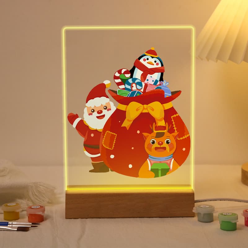 A 3D acrylic LED light box featuring a festive Christmas design with Santa Claus, a snowman, a reindeer, and a gift sack, glowing with a warm yellow light on a wooden base.