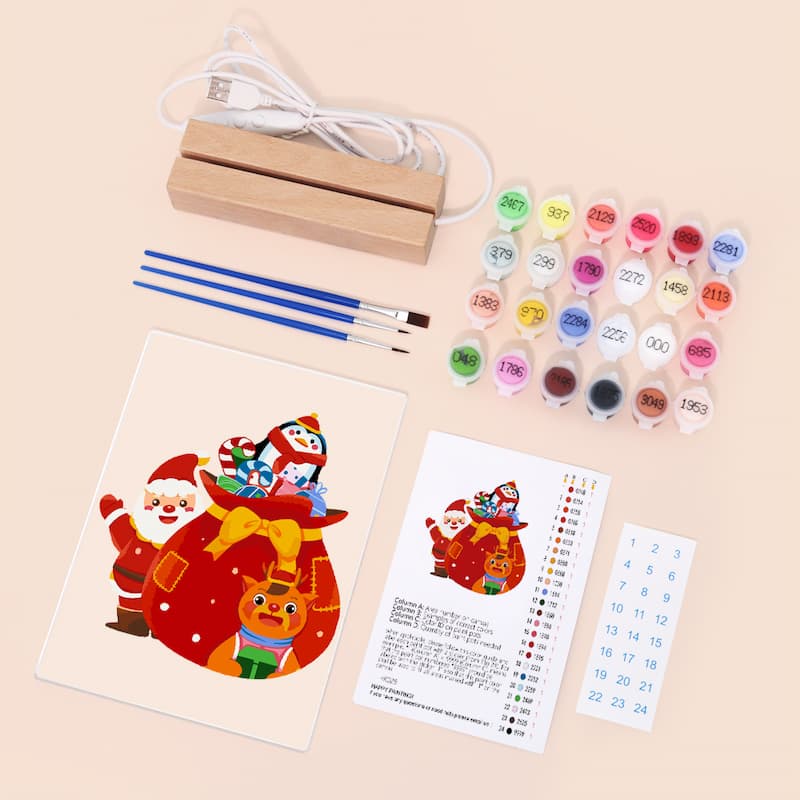 Christmas-themed DIY canvas painting kit laid out on a light beige background, including a printed design of Santa and a reindeer, paint tubes, small brushes, a wooden stand, and instruction sheets.