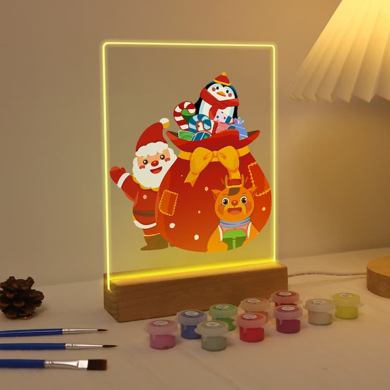 A 3D acrylic LED light box featuring a festive Christmas scene with Santa Claus, a penguin, a reindeer, and a gift sack, glowing with warm yellow light on a wooden base. Paint pots and paintbrushes are arranged on the table in front, with a lamp visible in the background.
