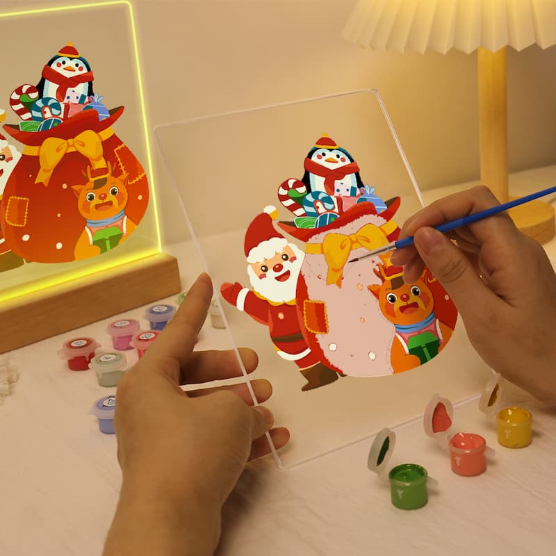 A person is painting a Christmas-themed acrylic panel with Santa Claus and a dog, holding a clear acrylic frame over a tabletop with small paint pots nearby. A finished glowing panel and a lamp are visible in the background.