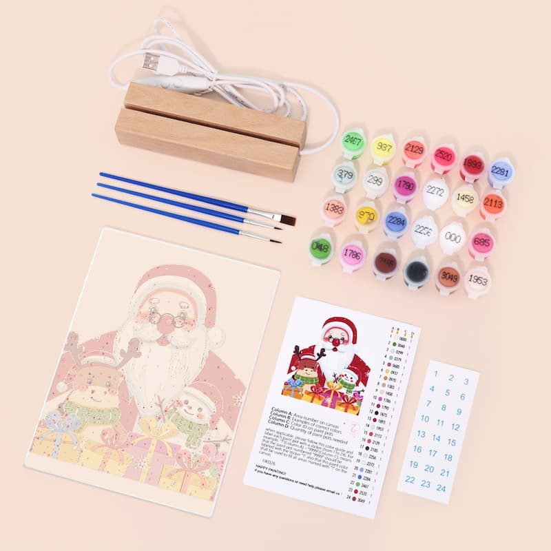 A Christmas-themed DIY craft kit laid out on a light beige background, including a printed Santa illustration, small paint pots with numbered caps, two blue-handled paintbrushes, a wooden stand, a USB charging cable, and an instruction sheet.