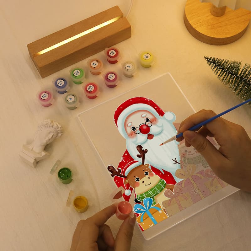 A person is painting a Christmas-themed canvas with Santa Claus and a reindeer using a small paintbrush, surrounded by paint pots, a wooden light box, and other craft supplies on a tabletop.
