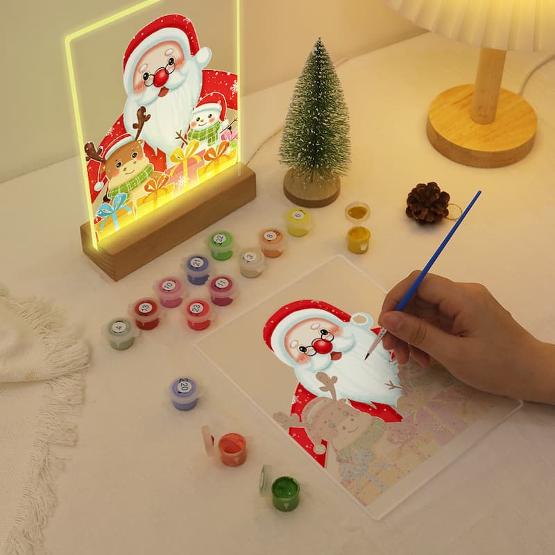 Decorative Christmas scene with a hand painting a Santa Claus design on paper, surrounded by small containers and a lit sign.
