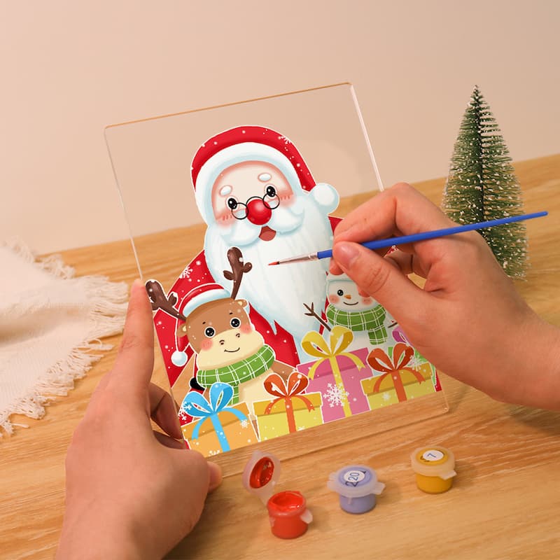 A person's hands are shown painting a colorful Christmas-themed acrylic panel with Santa Claus, a reindeer, and gifts. The scene is set on a wooden table with small paint pots and a miniature Christmas tree in the background.