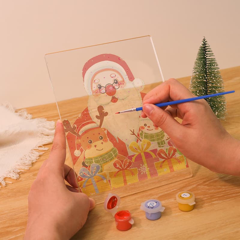 A person's hands are shown painting a clear acrylic panel with a Christmas design of Santa Claus, a reindeer, and snowman, using a fine-tip paintbrush. Small paint pots are visible on the wooden table next to a miniature Christmas tree.