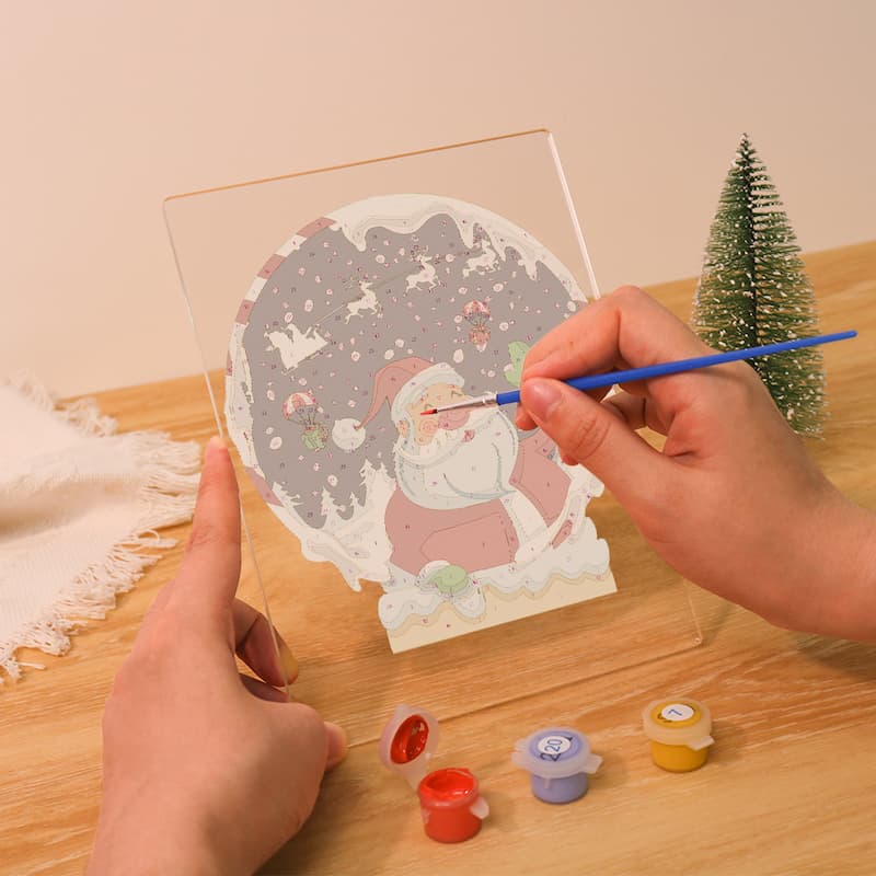 A person's hands are shown painting a circular Santa Claus winter scene on a clear acrylic panel, with small paint pots and a miniature Christmas tree on a wooden table.